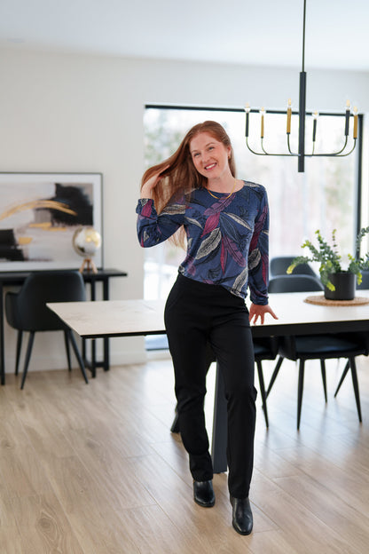 A woman wearing the St Charles Sweater by Rien ne se Perd in Foliage, a jewel toned patterned top with long sleeves and elastic cuffs. She is wearing it with the Murphy Pants in Black and standing in a modern kitchen. 
