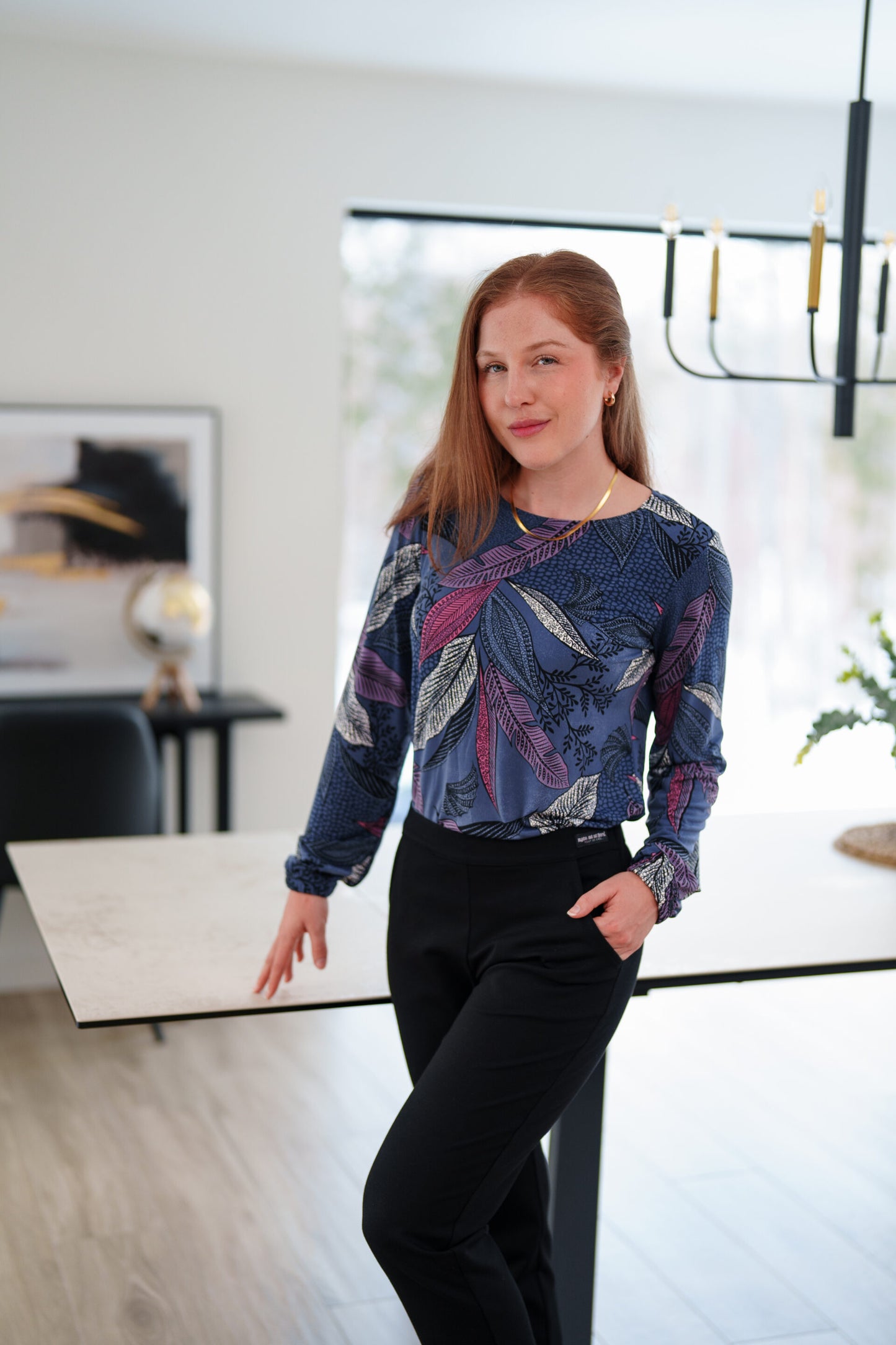 A woman wearing the St Charles Sweater by Rien ne se Perd in Foliage, a jewel toned patterned top with long sleeves and elastic cuffs. She is wearing it with the Murphy Pants in Black and standing in a modern kitchen. 
