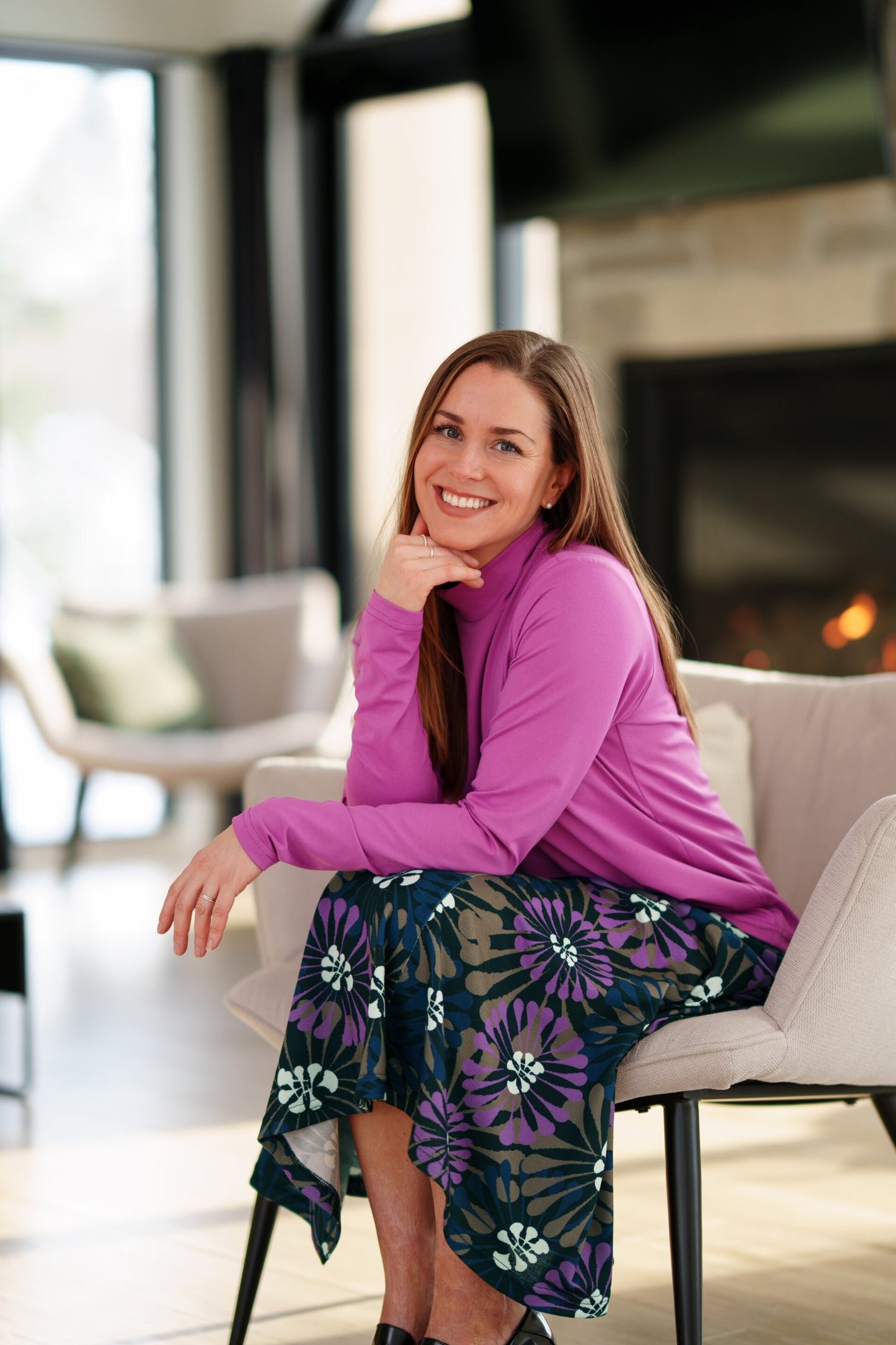 A woman wearing the Ste. Croix Sweater by Rien ne se Perd in Magenta, a mock turtleneck with long sleeves. She is wearing it with the Hudson Skirt in Floral print, and sitting in a light beige armchair in a living room setting with a stone fireplace