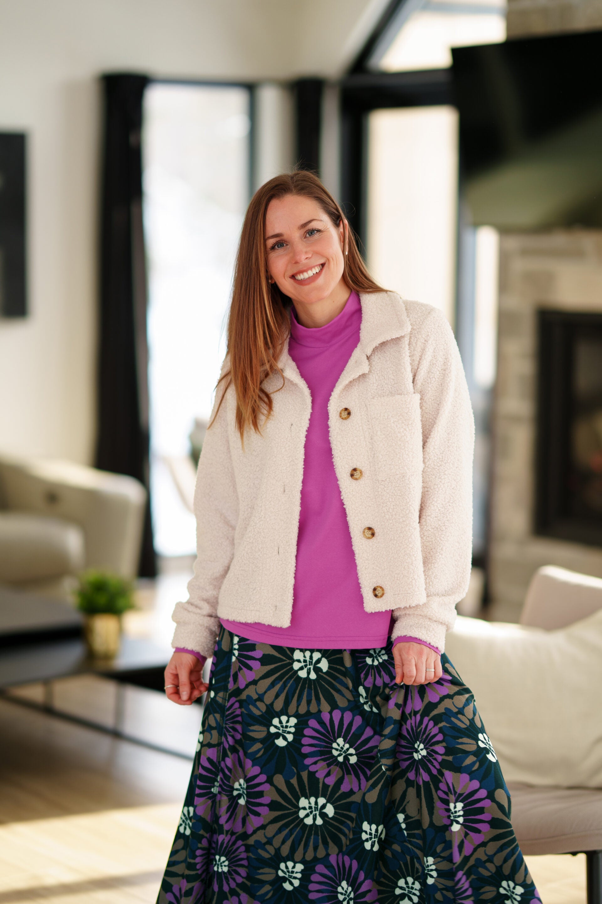 A woman wearing the Ste. Croix Sweater by Rien ne se Perd in Magenta, a mock turtleneck with long sleeves. She is wearing it with the Hudson Skirt in Floral print and a white boucle jacket and standing in a living room. 