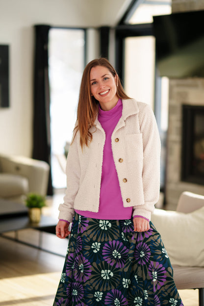 A woman wearing the Ste. Croix Sweater by Rien ne se Perd in Magenta, a mock turtleneck with long sleeves. She is wearing it with the Hudson Skirt in Floral print and a white boucle jacket and standing in a living room. 