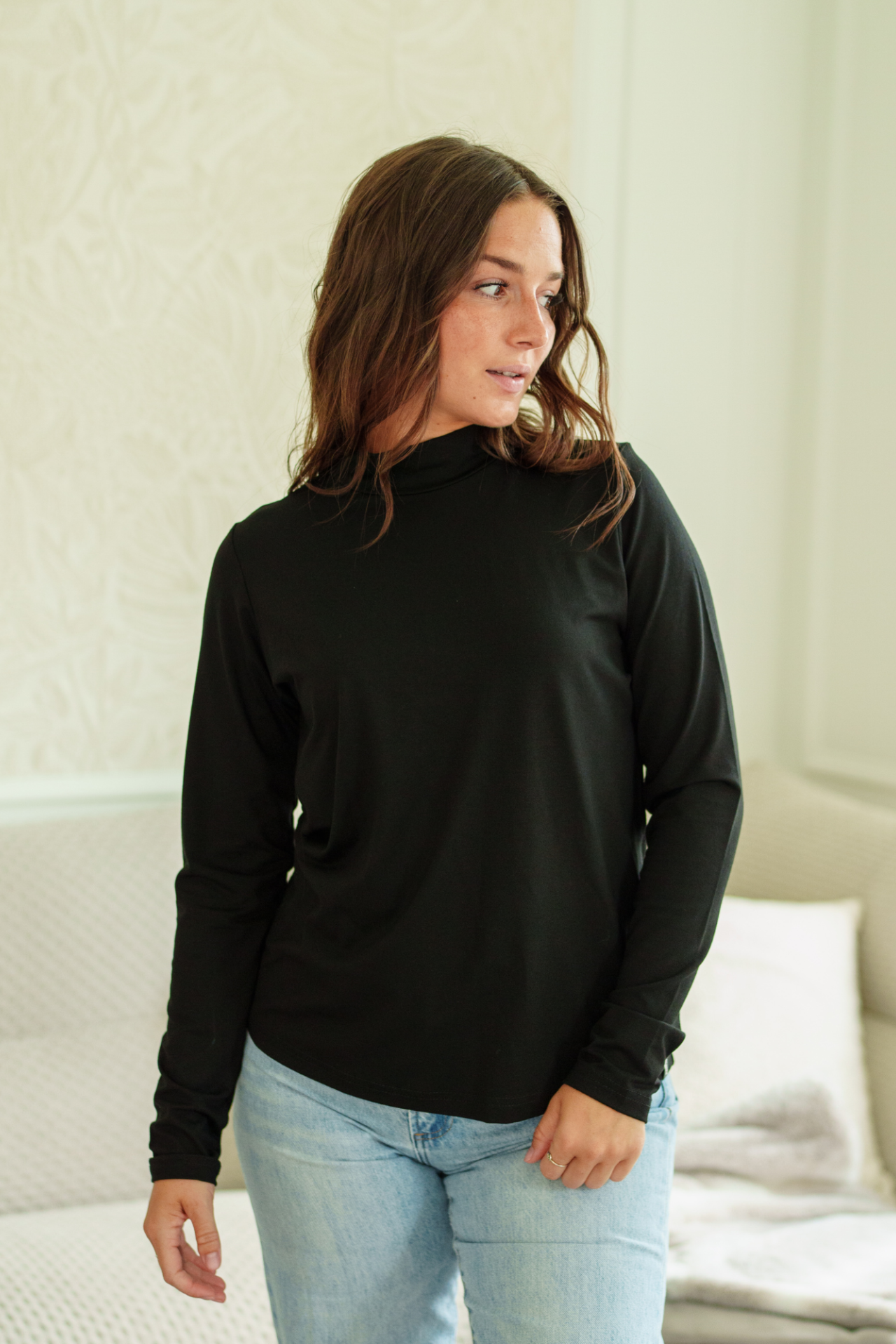 A woman wearing the Ste. Croix Sweater by Rien ne se Perd in Black, a  long-sleeve mock neck top. She is wearing it with light blue jeans, standing in a brightly lit living room. 