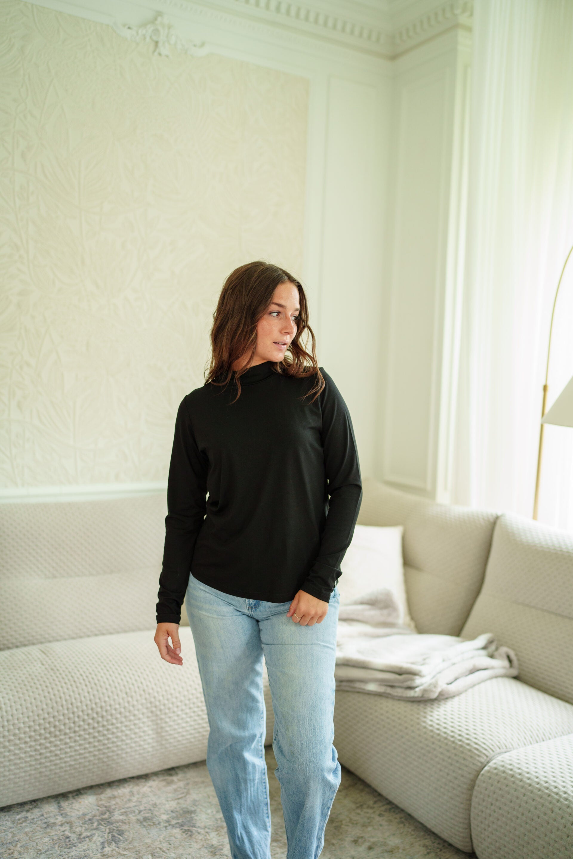 A woman wearing the Ste. Croix Sweater by Rien ne se Perd in Black, a  long-sleeve mock neck top. She is wearing it with light blue jeans, standing in a brightly lit living room. 