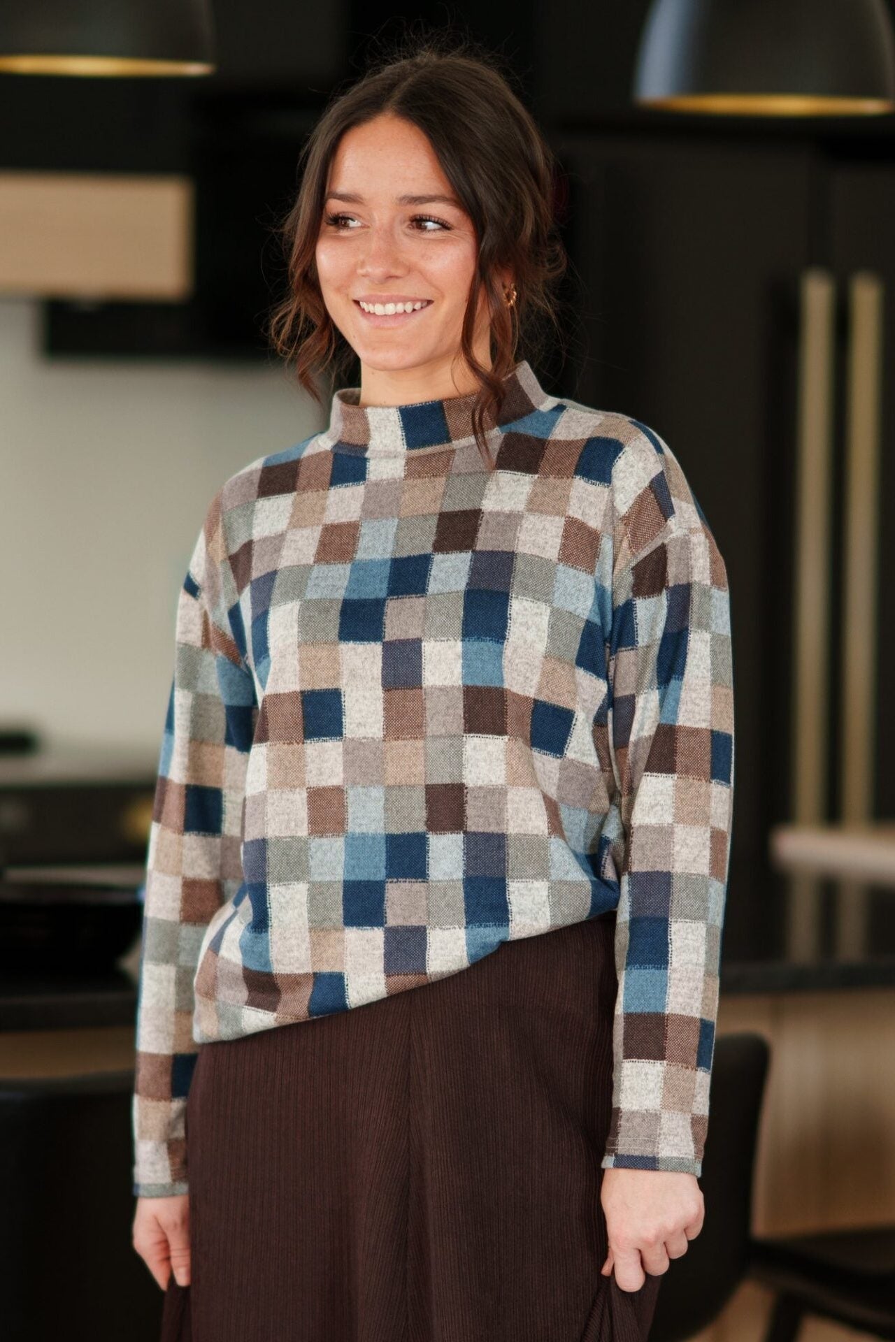 A woman wearing the Leran Sweater by Rien ne se Perd in Checkered print, a mock-neck knit sweater with a blue, brown, and beige pattern. She is wearig it with a brown skirt and standing in a modern kitchen setting. 