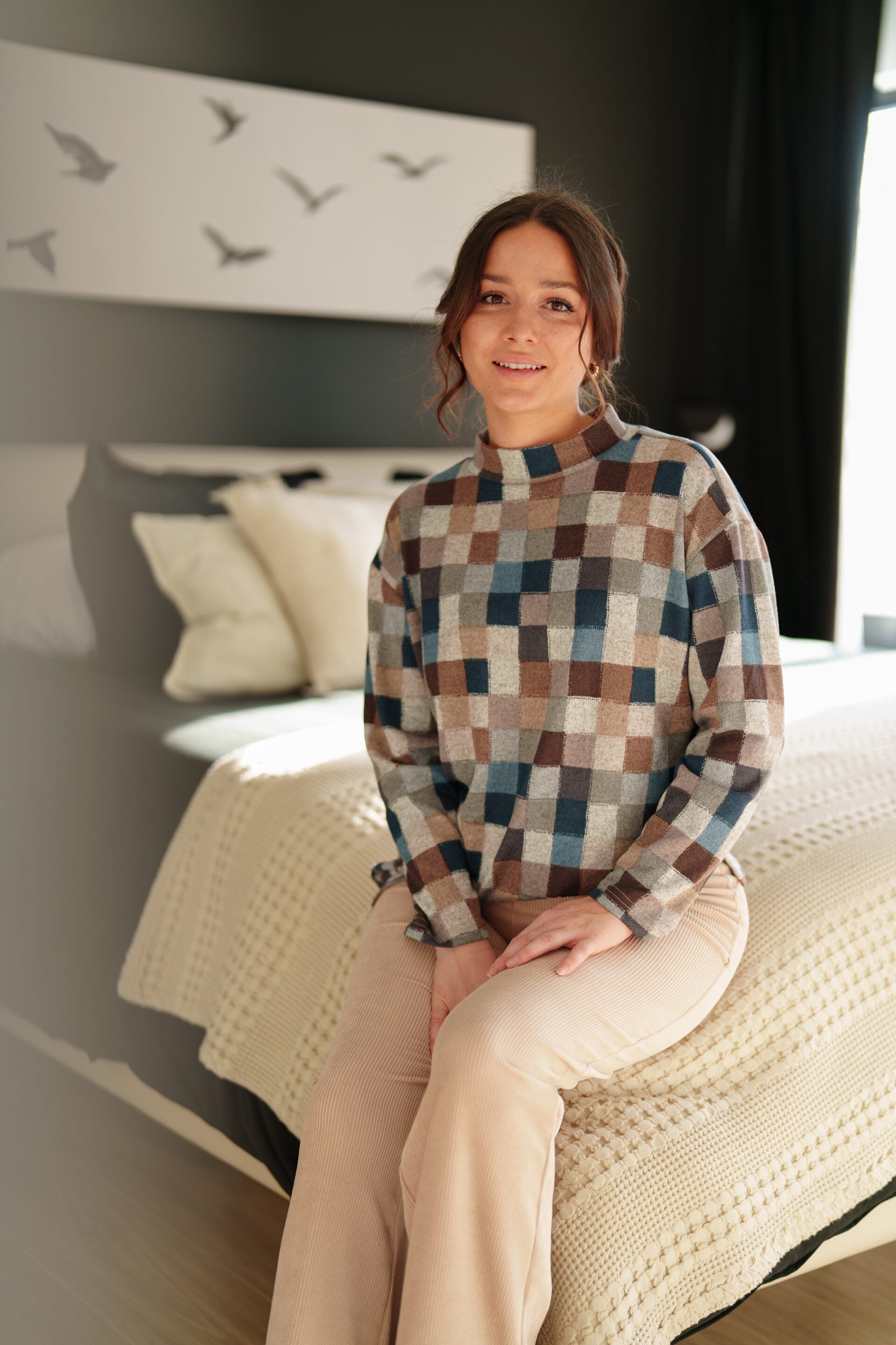 A woman wearing the Leran Sweater by Rien ne se Perd in Checkered print, a mock-neck knit sweater with a blue, brown, and beige pattern. She is wearing it with beige pants and sitting on a bed. 