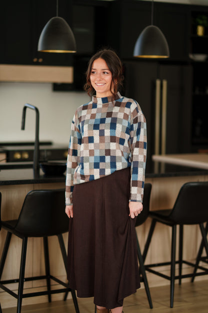 A woman wearing the Leran Sweater by Rien ne se Perd in Checkered print, a mock-neck knit sweater with a blue, brown, and beige pattern. She is wearig it with a brown skirt and standing in a modern kitchen setting. 