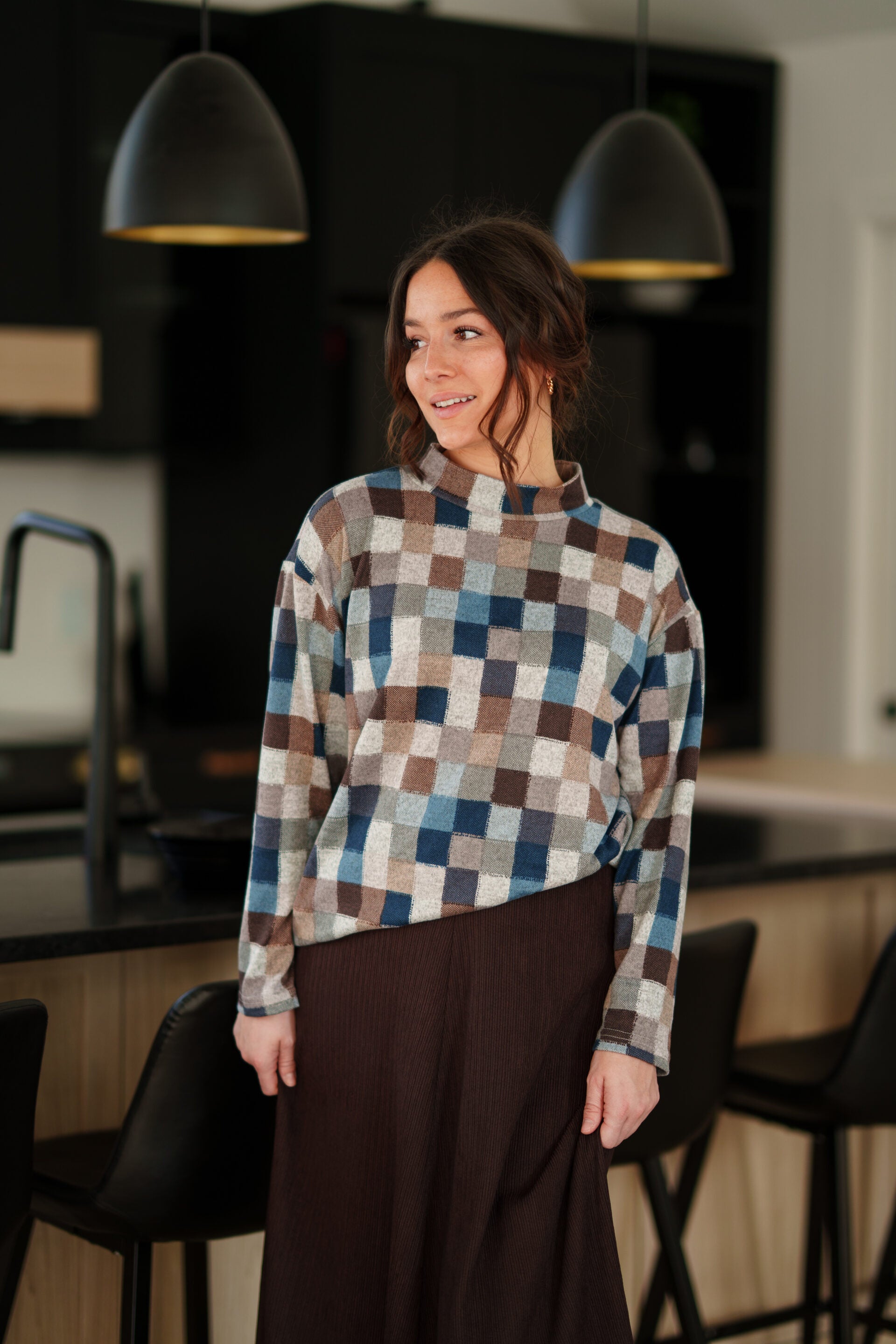 A woman wearing the Leran Sweater by Rien ne se Perd in Checkered print, a mock-neck knit sweater with a blue, brown, and beige pattern. She is wearig it with a brown skirt and standing in a modern kitchen setting. 