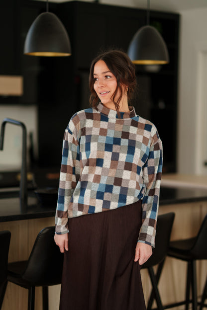 A woman wearing the Leran Sweater by Rien ne se Perd in Checkered print, a mock-neck knit sweater with a blue, brown, and beige pattern. She is wearig it with a brown skirt and standing in a modern kitchen setting. 