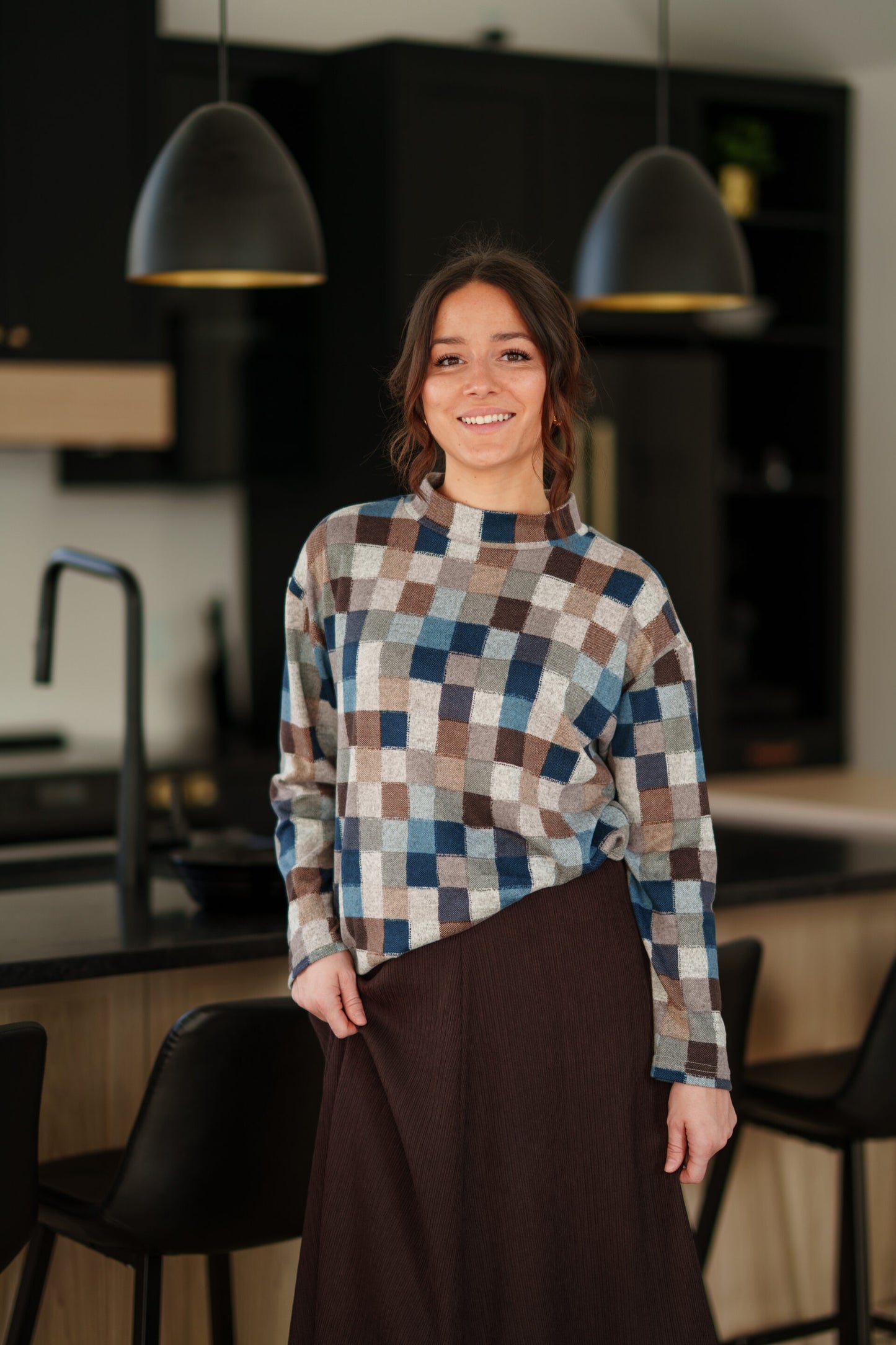 A woman wearing the Leran Sweater by Rien ne se Perd in Checkered print, a mock-neck knit sweater with a blue, brown, and beige pattern. She is wearig it with a brown skirt and standing in a modern kitchen setting. 