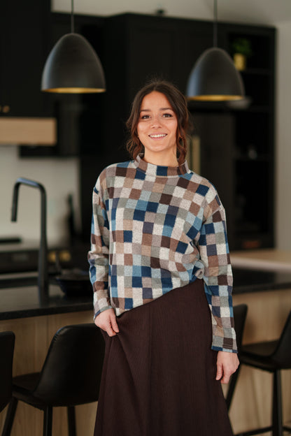 A woman wearing the Leran Sweater by Rien ne se Perd in Checkered print, a mock-neck knit sweater with a blue, brown, and beige pattern. She is wearig it with a brown skirt and standing in a modern kitchen setting. 