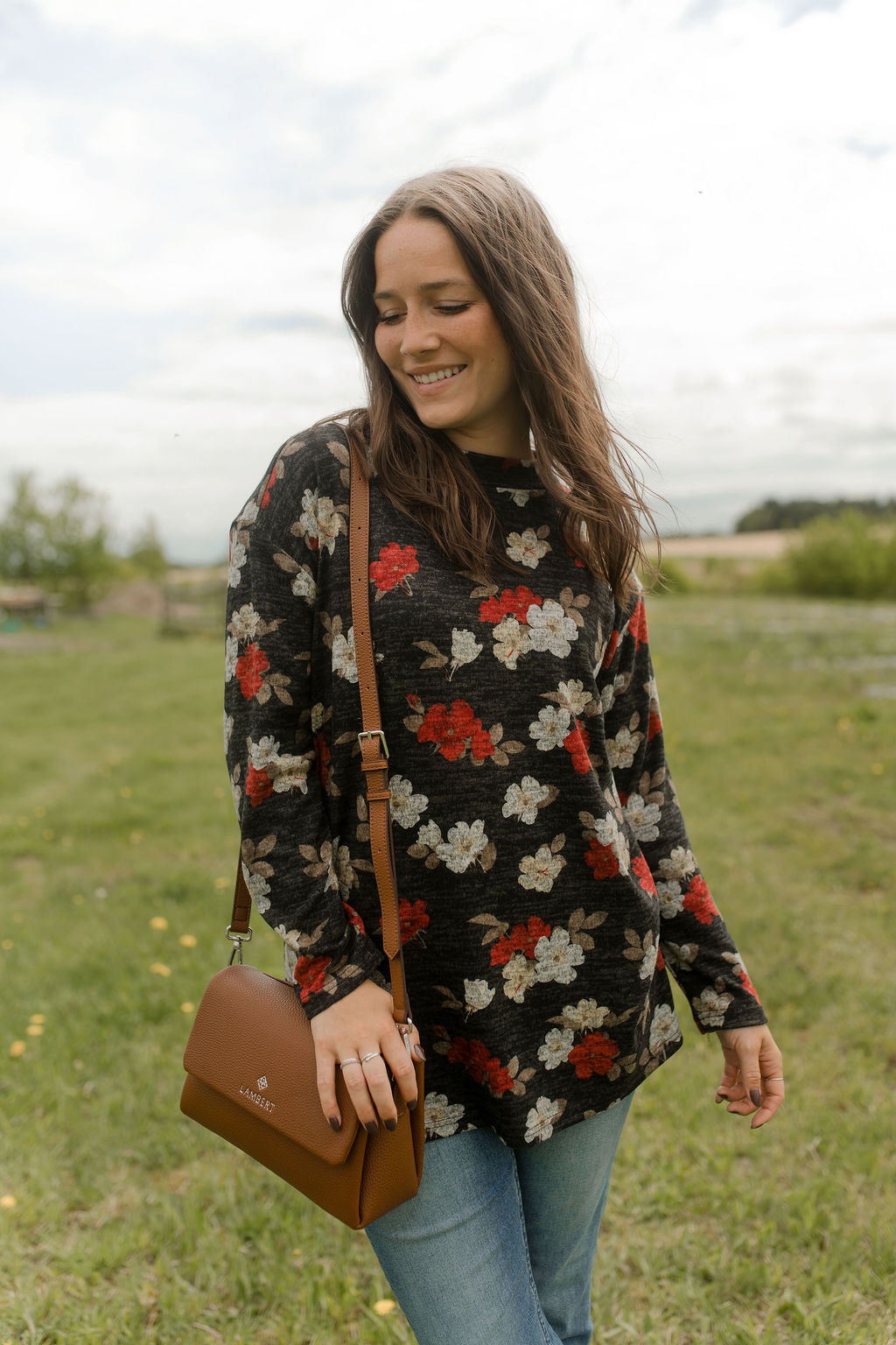 A woman wearing the Leran Sweater by Rien ne se Perd in Floral print, a mock-neck knit sweater with a black, white and red pattern. She is wearing it with jeans and standing outside in a field. 