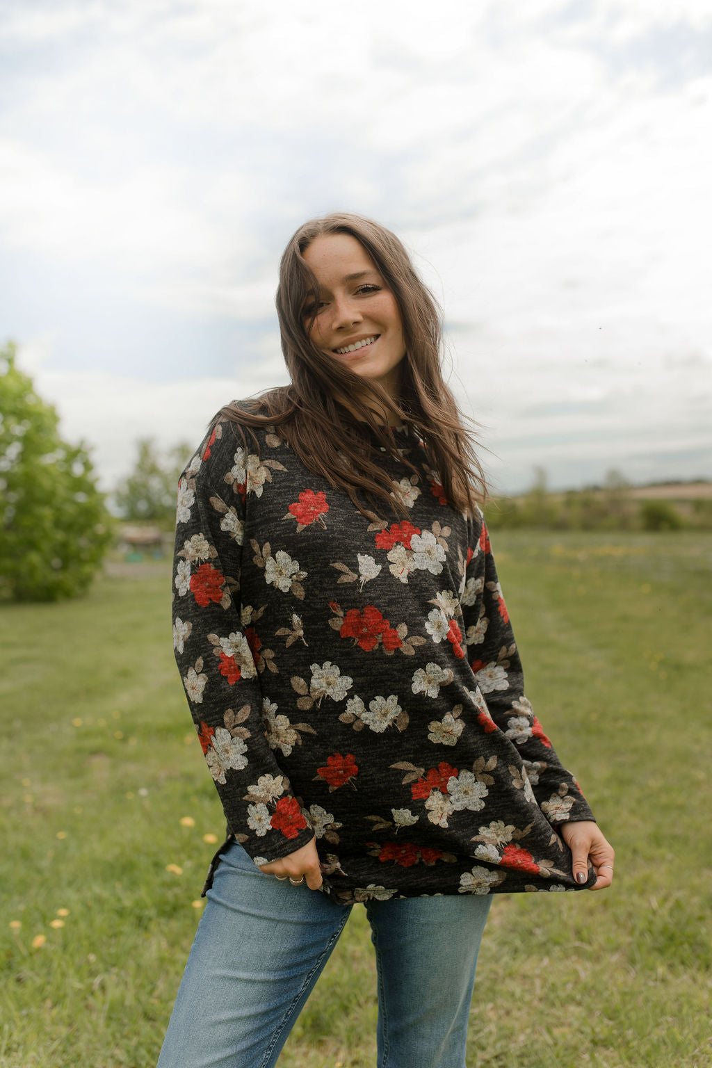 A woman wearing the Leran Sweater by Rien ne se Perd in Floral print, a mock-neck knit sweater with a black, white and red pattern. She is wearing it with jeans and standing outside in a field. 