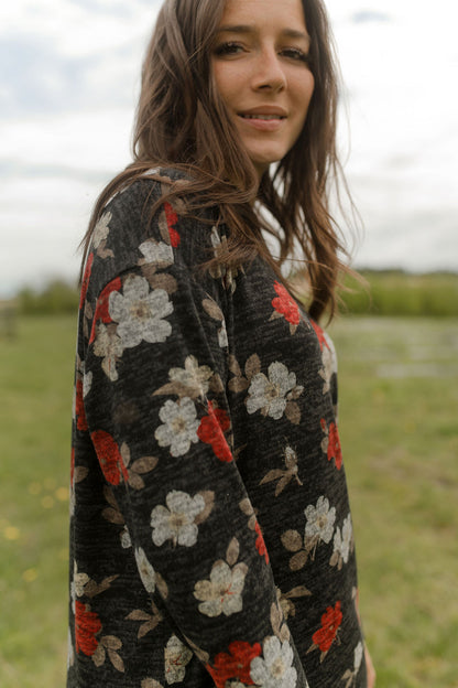 Side view of a woman wearing the Leran Sweater by Rien ne se Perd in Floral print, a mock-neck knit sweater with a black, white and red pattern. She is wearing it with jeans and standing outside in a field. 