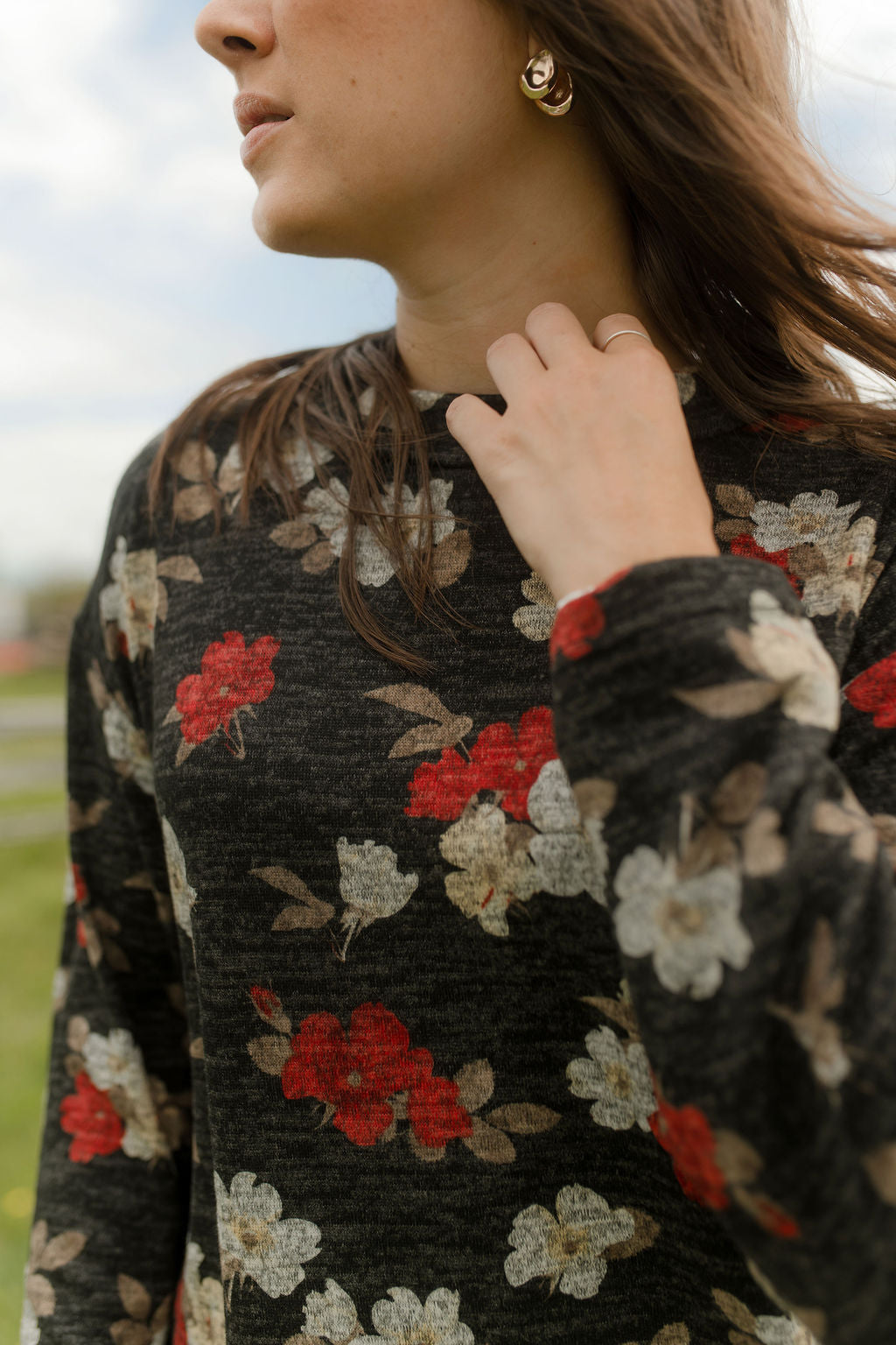 Close up of a woman wearing the Leran Sweater by Rien ne se Perd in Floral print, a mock-neck knit sweater with a black, white and red pattern. She is wearing it with jeans and standing outside in a field. 
