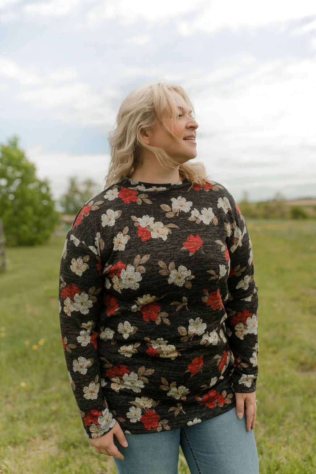 A woman wearing the Leran Sweater by Rien ne se Perd in Floral print, a mock-neck knit sweater with a black, white and red pattern. She is wearing it with jeans and standing outside in a field. 