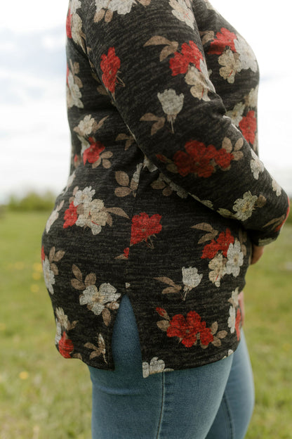 Side view of a woman wearing the Leran Sweater by Rien ne se Perd in Floral print, a mock-neck knit sweater with a black, white and red pattern. She is wearing it with jeans and standing outside in a field. 