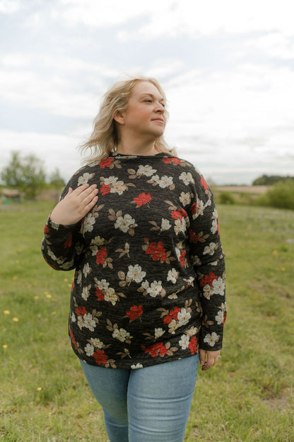 A woman wearing the Leran Sweater by Rien ne se Perd in Floral print, a mock-neck knit sweater with a black, white and red pattern. She is wearing it with jeans and standing outside in a field. 