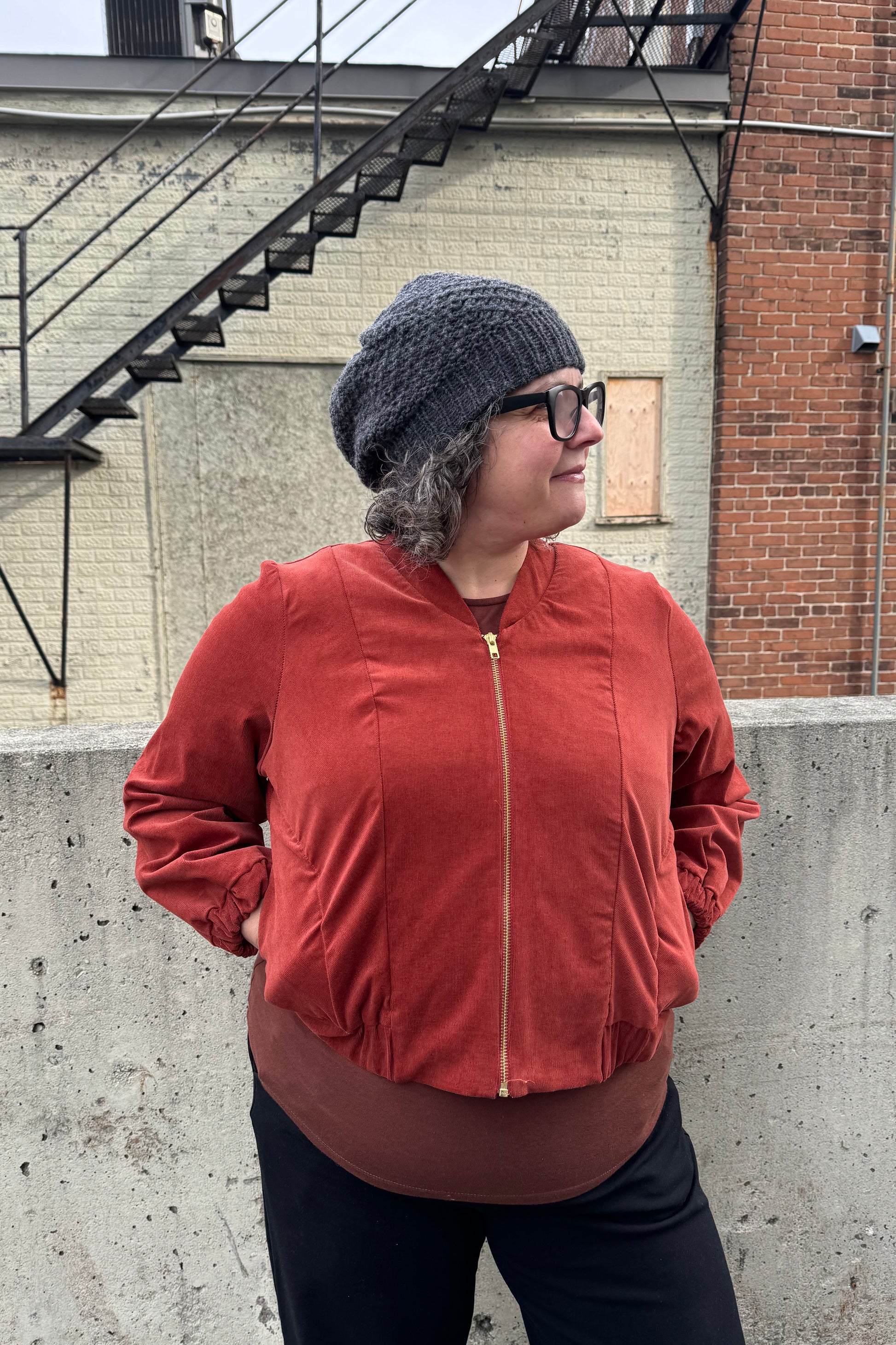 A woman wearing the Beret by Charlevoix Pure Laine in Charcoal, with the Redrawn Zip Jacket by Eve Lavoie in Terracotta Corduroy. She is standing outside. 