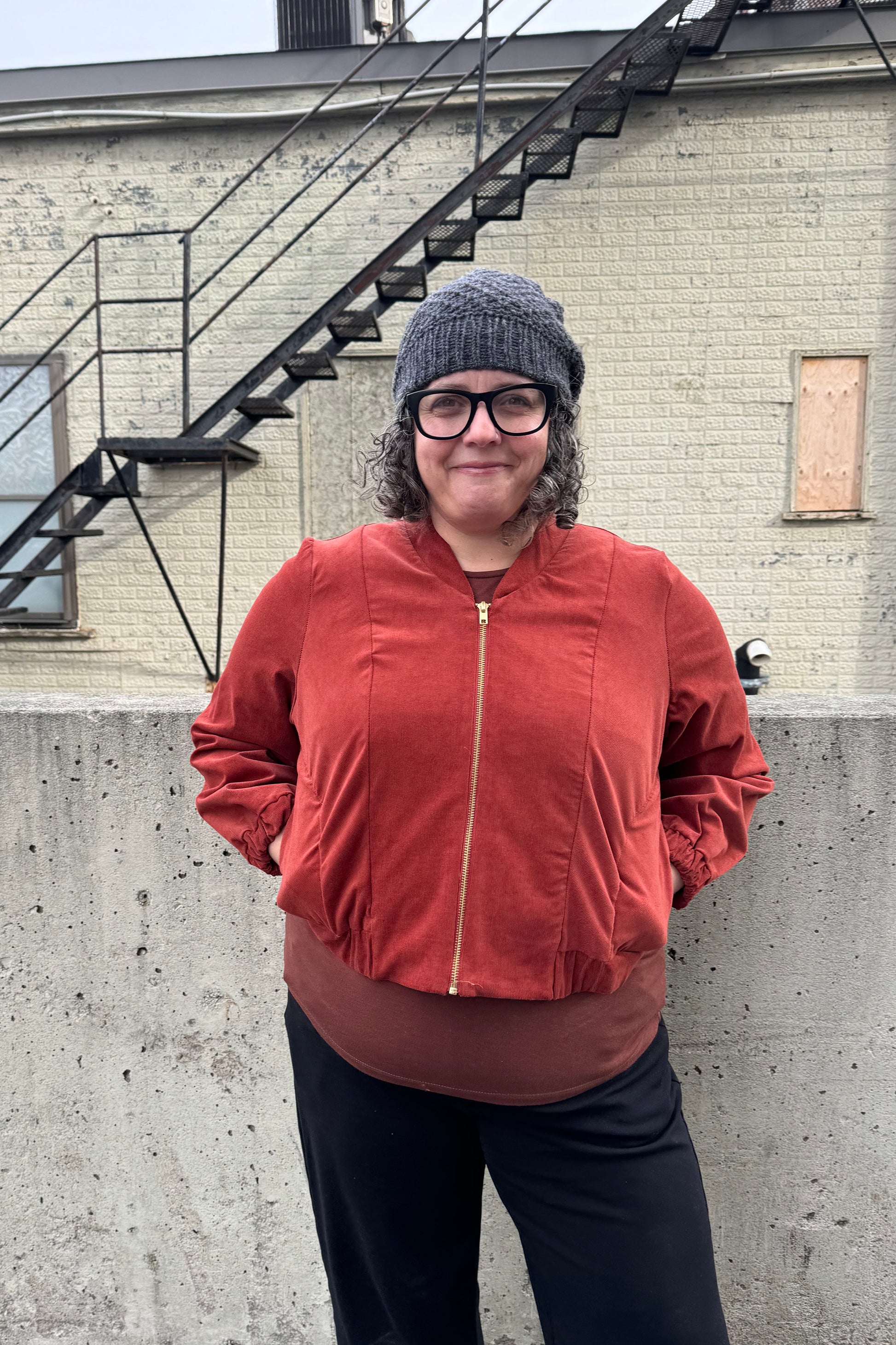 A woman wearing the Beret by Charlevoix Pure Laine in Charcoal, with the Redrawn Zip Jacket by Eve Lavoie in Terracotta Corduroy. She is standing outside. 