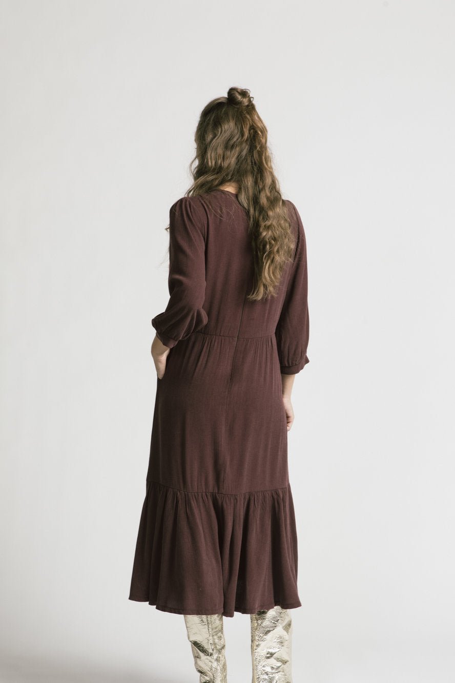 Back view of a woman wearing the Chatterley Dress by Allison Wonderland in Mulberry, standing in front of a white background 