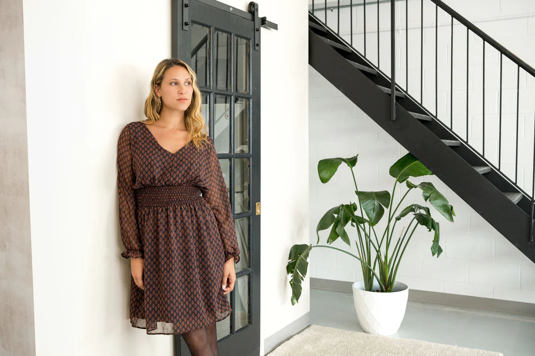 A woman wearing the Spritz Dress by Cherry Bobin in Brown Pattern, featuring puff sleeves, a v-neck,  cinch waisted, and a full knee-length skirt. She is standing in a white room with a black staircase. 