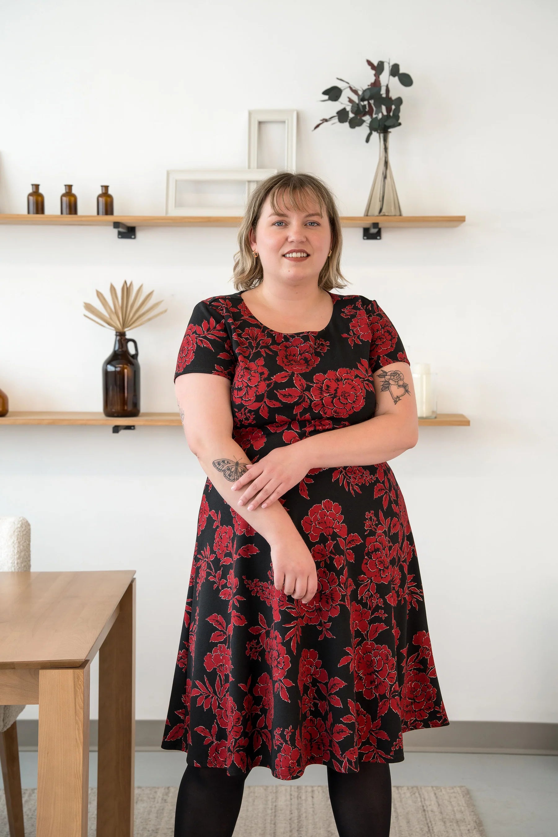 A woman wearing the Bellini Dress by Cherry Bobin in Red Floral, with short sleeves, a round neck, and a fit and flare shape that falls to the knee. She is standing in a room with  a wooden table. 