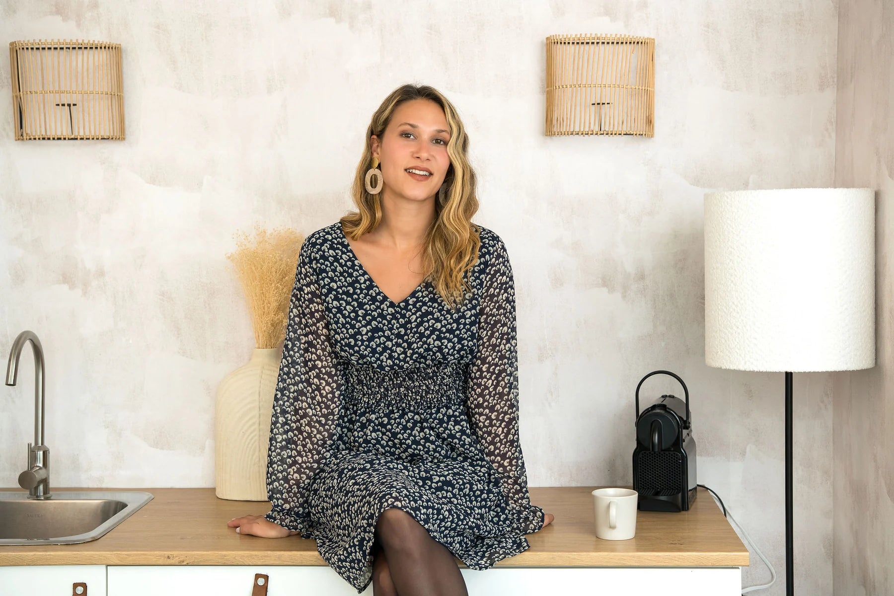A woman wearing the Spritz Dress by Cherry Bobin in Blue Pattern, featuring puff sleeves, a v-neck,  cinch waisted, and a full knee-length skirt. She is sitting on a wooden counter. 
