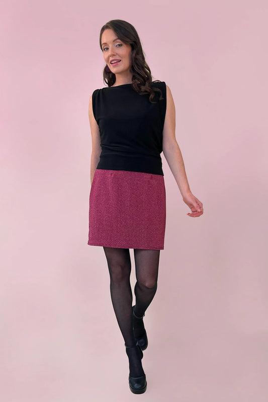 A woman wearing the Glitter Skirt by Canopée in Pink, a short stretchy skirt She's wearing it with a black sleeveless top against a pink background.