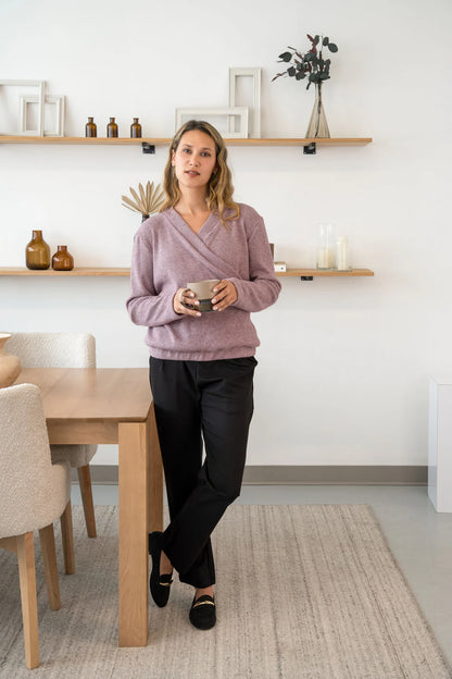 A woman wearing the Mimosa Sweater by Cherry Bobin in Pink, a chunky knit sweater with a faux-wrap neckline, long slightly puffed sleeves, and an elastic waistband. She is wearing it with black pants and holding a mug while standing by a table. 