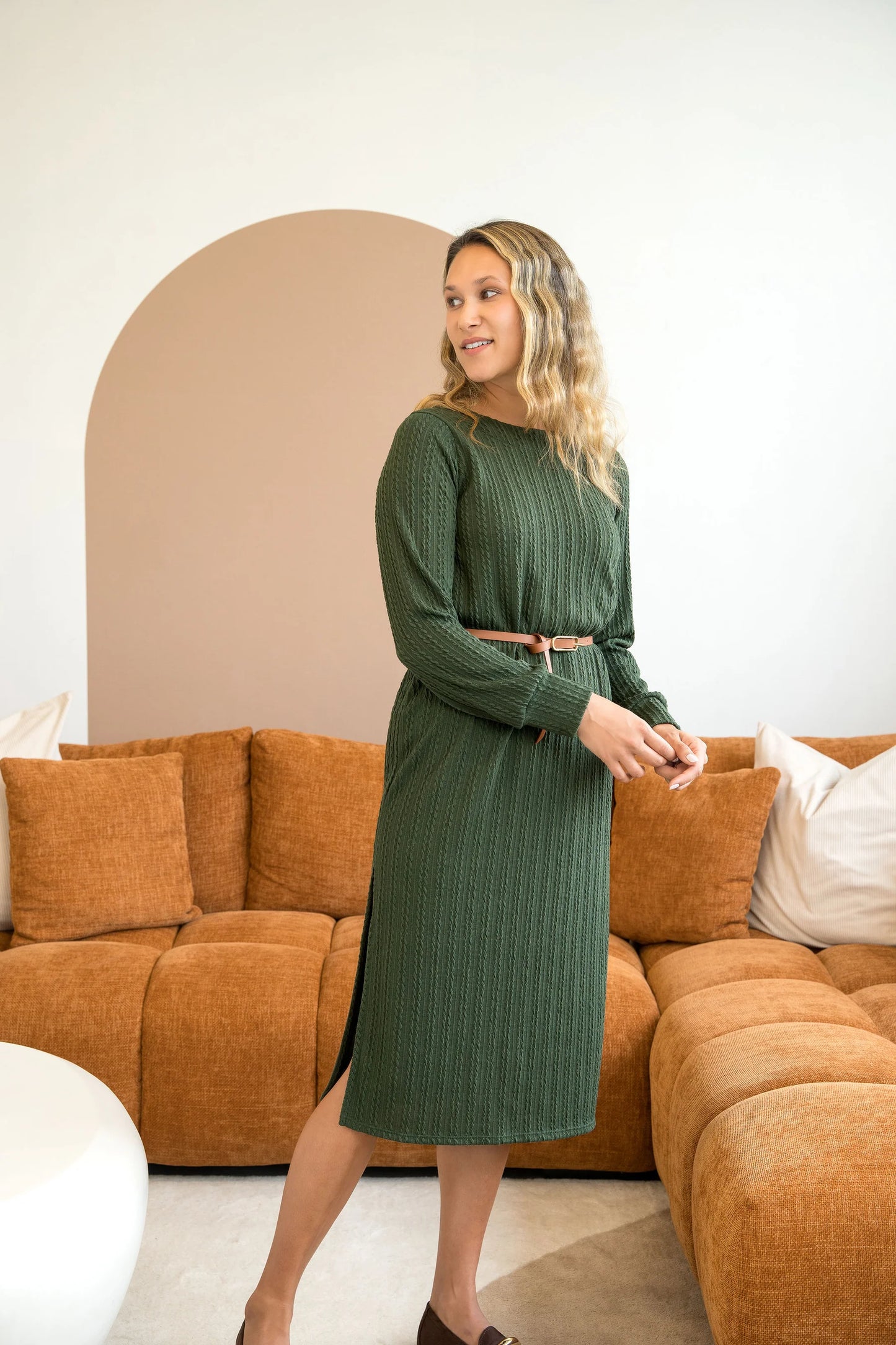 A woman wearing the Negroni Dress by Cherry Bobin in Green, a mid-calf dress with in cable-knit textured fabric, side slits, and long, slighty puffed sleeves. She is standing in a room with a brown couch. 