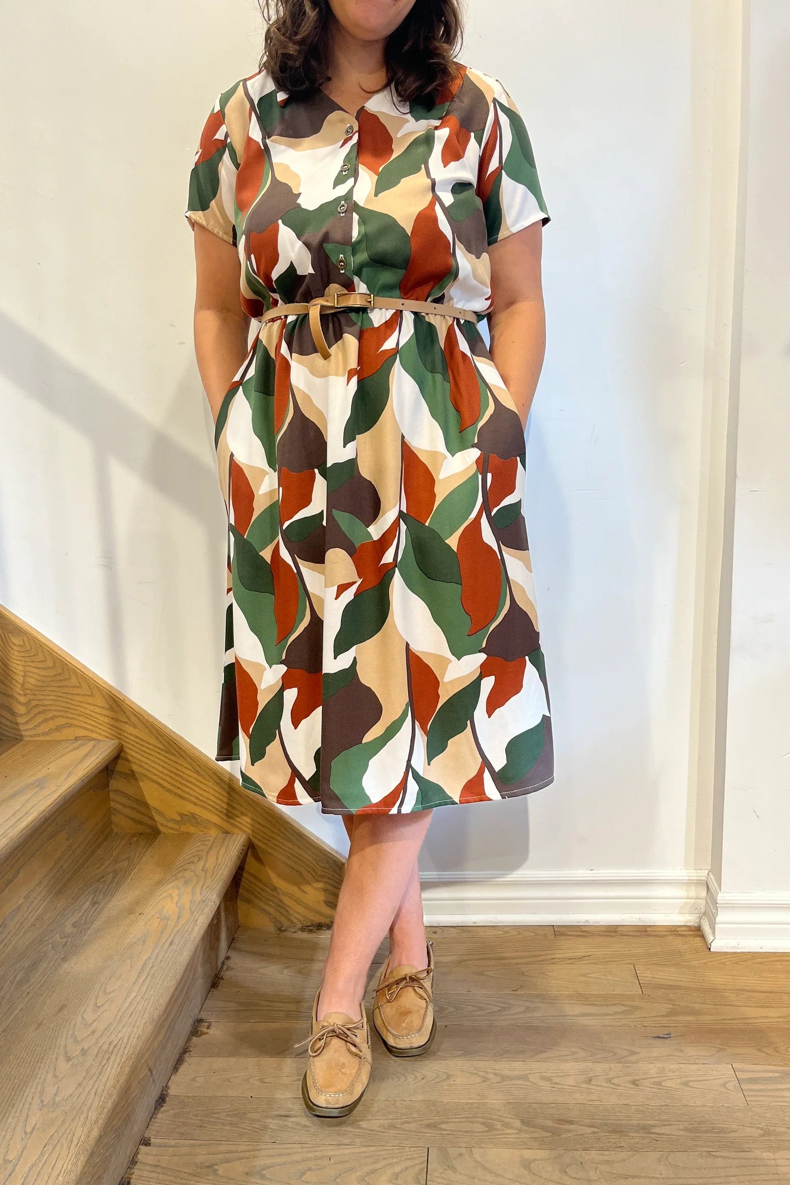 Neck down view of a woman wearing the Paloma Dress by Cherry Bobin in Brown Green Ivory, with a classic collar, vintage brass buttons, short sleeves, an elastic waist, and a full below-the-knee skirt. She is standing by a staircase. 