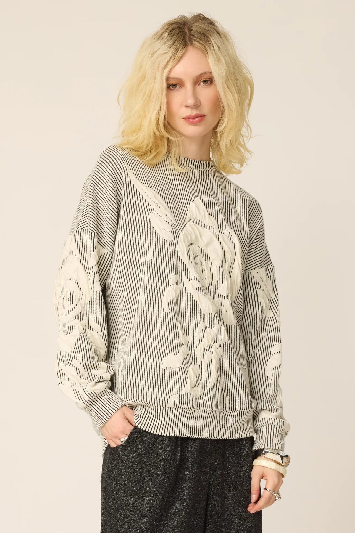 A woman wearing the Howard Sweater from Chlo by Cokluch in Natural, with a striped fabric and floral applique in an oversized cut. She is wearing it with grey pants and standing againts a beige background