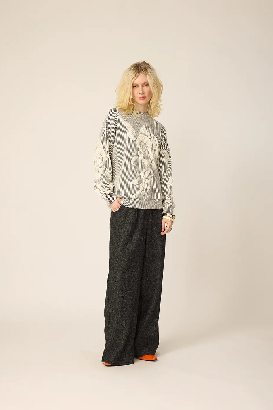 A woman wearing the Howard Sweater from Chlo by Cokluch in Natural, with a striped fabric and floral applique in an oversized cut. She is wearing it with grey pants and standing againts a beige background