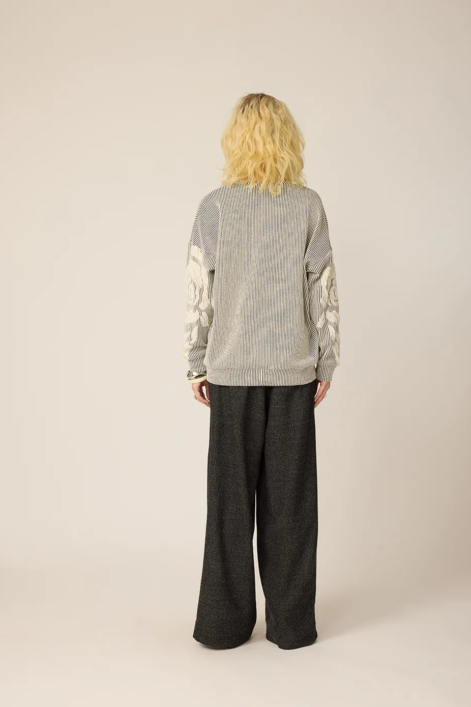 Back view of a woman wearing the Howard Sweater from Chlo by Cokluch in Natural, with a striped fabric and floral applique in an oversized cut. She is wearing it with grey pants and standing againts a beige background