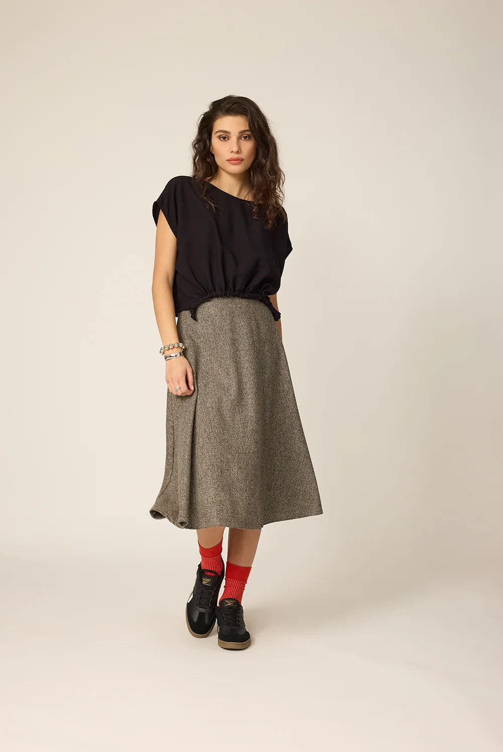 A woman wearing the Amber Skirt from Chlo by Cokluch in Greige Tweed, a full, flared mid-calf skirt with a high waist and adjustable side drawstrings with metal rings.  She is wearing it with a black top and standing against a grey background. 