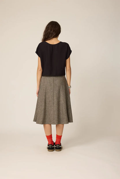 Back view of a woman wearing the Amber Skirt from Chlo by Cokluch in Greige Tweed, a full, flared mid-calf skirt with a high waist and adjustable side drawstrings with metal rings.  She is wearing it with a black top and standing against a grey background.