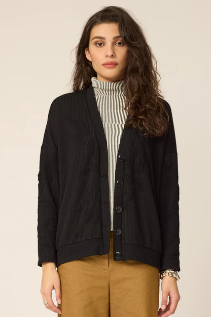 Woman wearing the Gus Cardigan from Cokluch in Black with ribbed fabric and floral embellishments on the front and sleeves. It has a relaxed it and five buttons. She is wearing it over an a striped top and ochre pants in front of a white background.