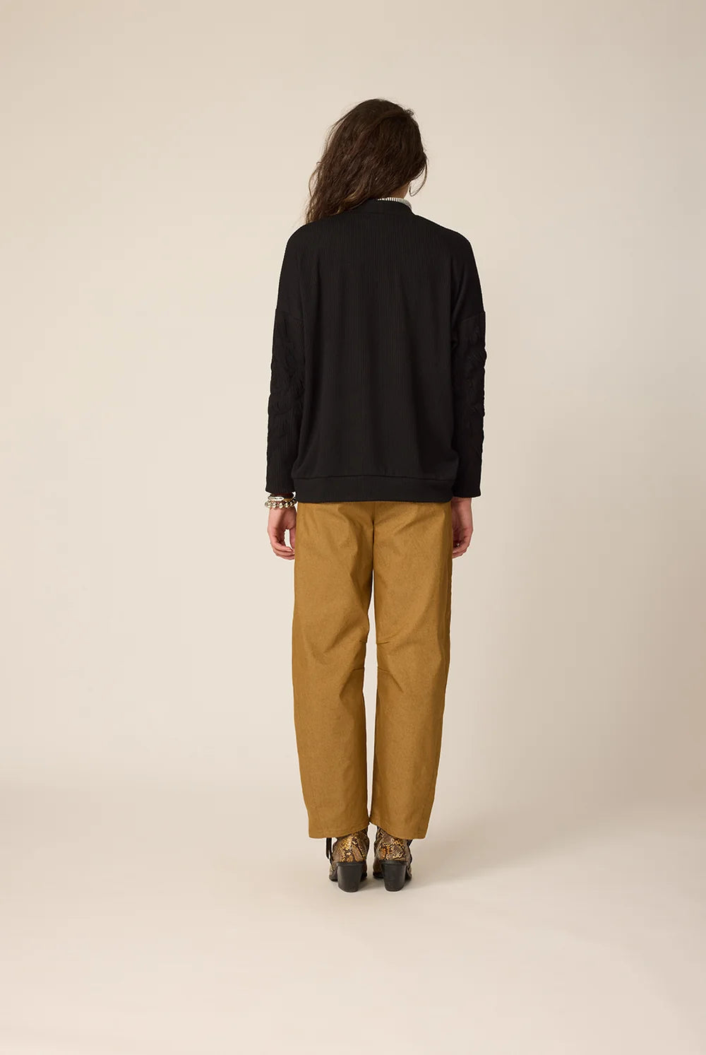 Back view of a woman wearing the Gus Cardigan from Cokluch in Black with ribbed fabric and floral embellishments on the front and sleeves. It has a relaxed it and five buttons. She is wearing it over an a striped top and ochre pants in front of a white background.