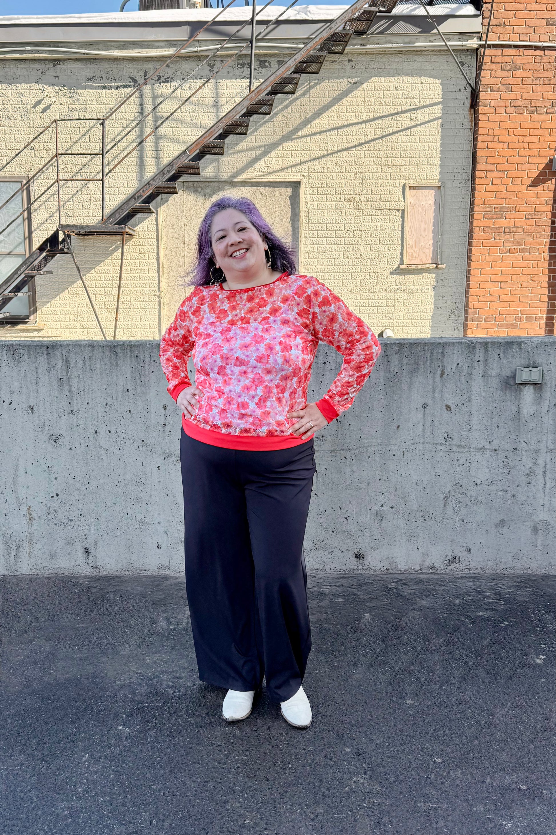 A woman wearing the Daisy Lace Shirt by Karkass in Coral, a floral lace shirt with solid cuffs and hem. She is wearing it with black pants and standing outside by a brick wall. 