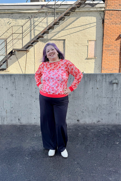 A woman wearing the Daisy Lace Shirt by Karkass in Coral, a floral lace shirt with solid cuffs and hem. She is wearing it with black pants and standing outside by a brick wall. 