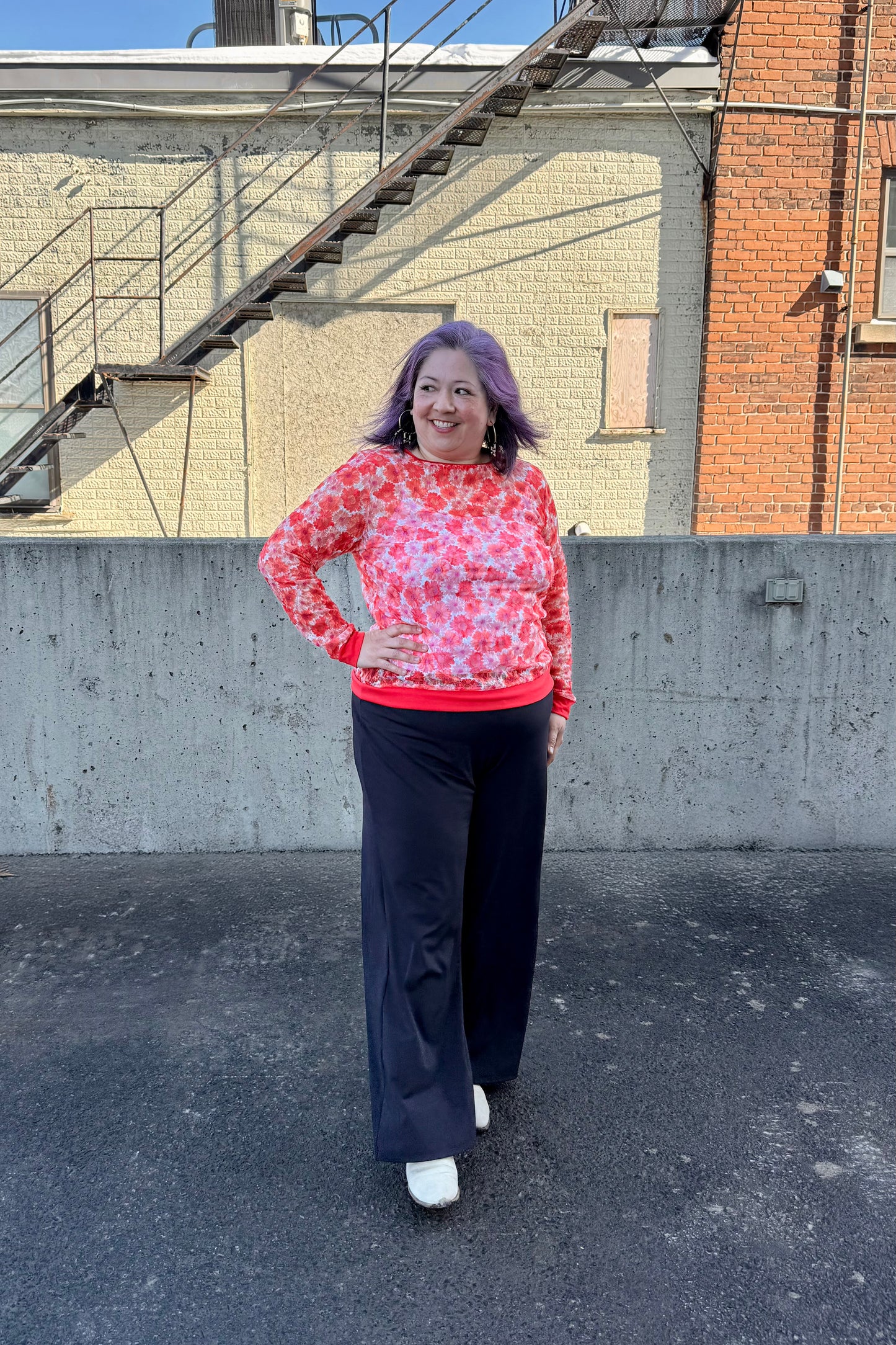 A woman wearing the Daisy Lace Shirt by Karkass in Coral, a floral lace shirt with solid cuffs and hem. She is wearing it with black pants and standing outside by a brick wall. 