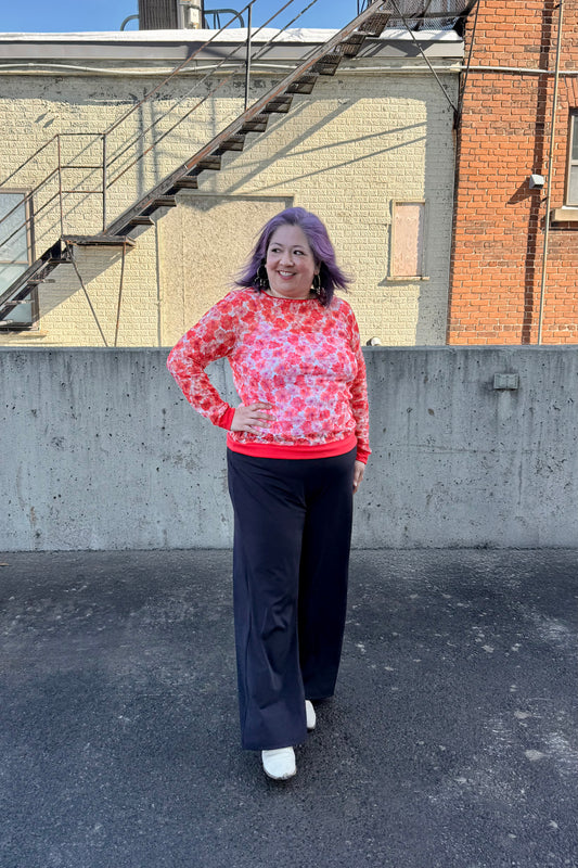 A woman wearing the Daisy Lace Shirt by Karkass in Coral, a floral lace shirt with solid cuffs and hem. She is wearing it with black pants and standing outside by a brick wall. 