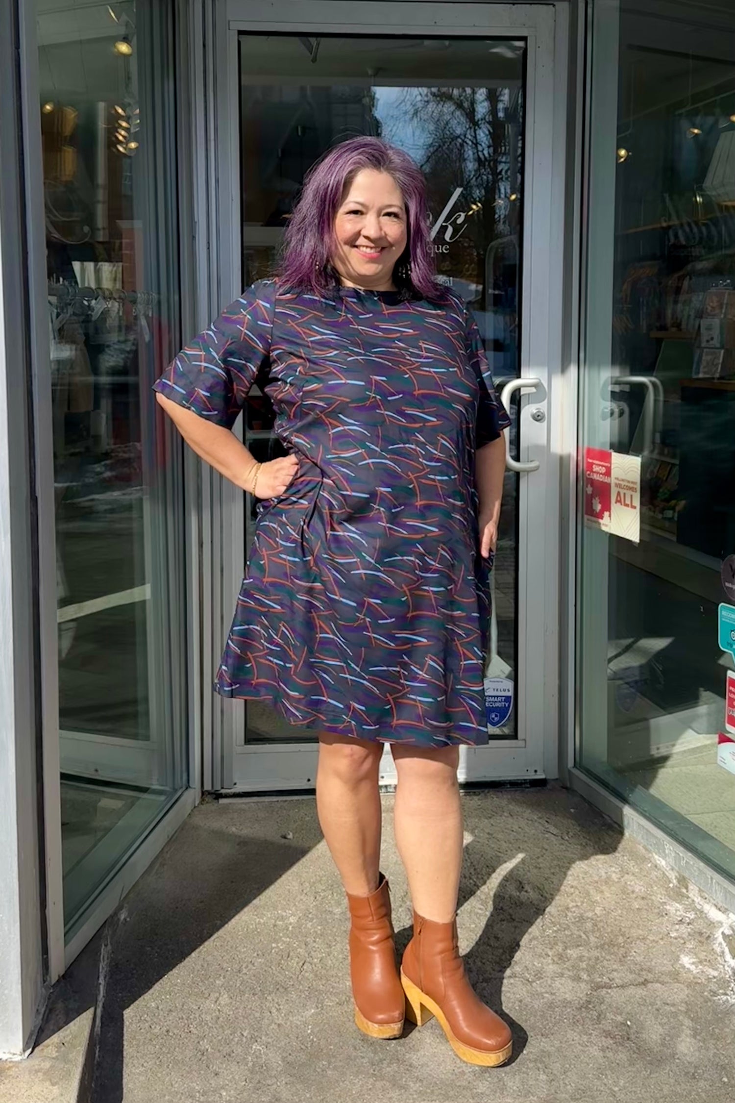Christina is wearing the Cecily Simple Shift Dress from Mandala in Dark Stix print. It has a high neck, short sleeves, a knee-length hem, French darts, and elastic at the back of the waist. She is standing outside of Flock. 