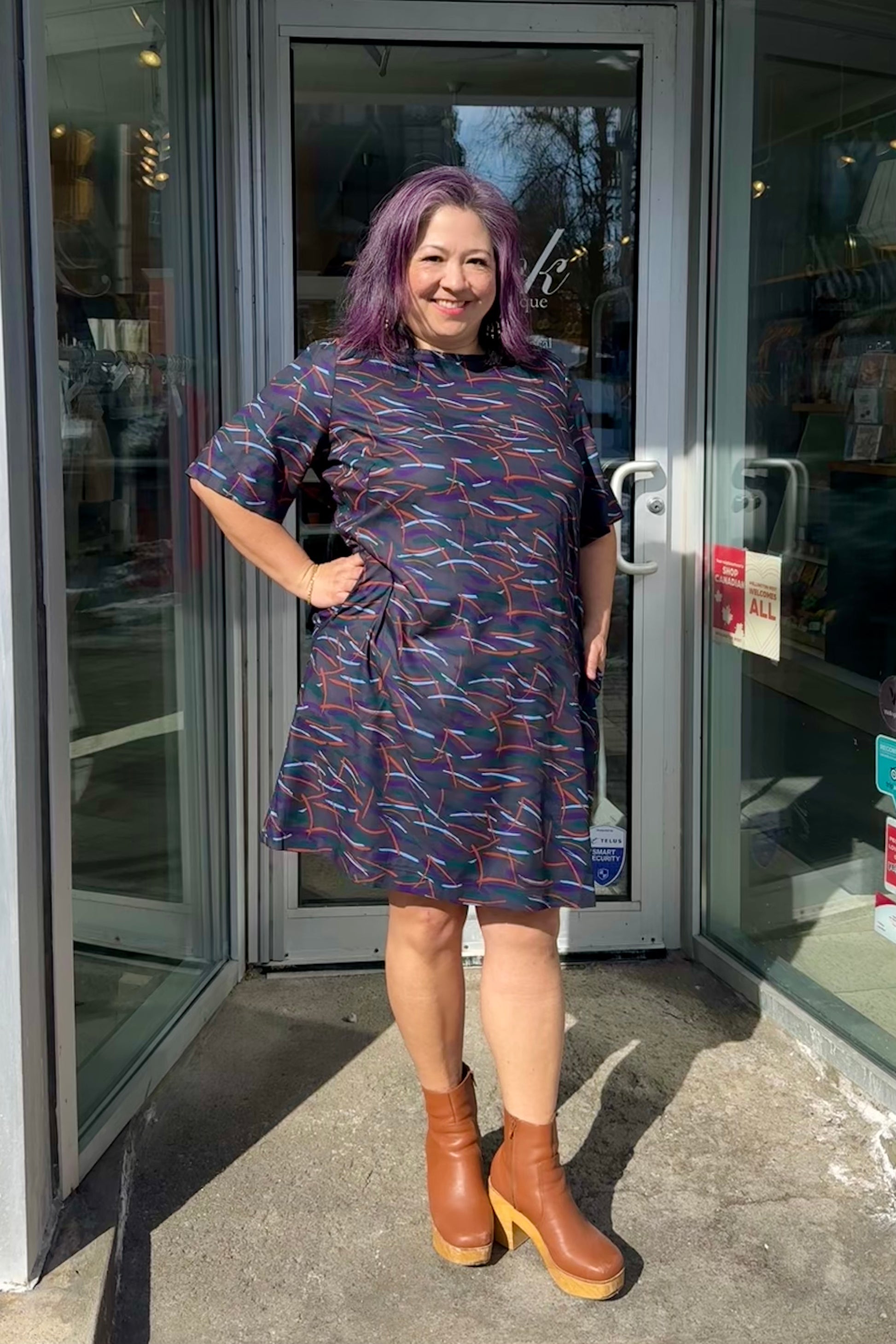 Christina is wearing the Cecily Simple Shift Dress from Mandala in Dark Stix print. It has a high neck, short sleeves, a knee-length hem, French darts, and elastic at the back of the waist. She is standing outside of Flock. 