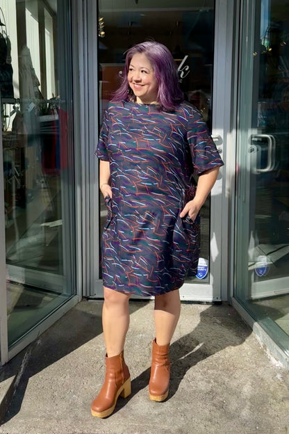 Christina is wearing the Cecily Simple Shift Dress from Mandala in Dark Stix print. It has a high neck, short sleeves, a knee-length hem, French darts, and elastic at the back of the waist. She is standing outside of Flock. 