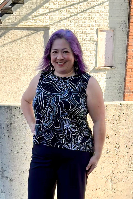 A woman wearing the Sunrise Camisole by Rien ne se Perd in Black/White floral, with a wide neck, wide straps, and a rounded hi-low hemline. She is wearing it with black pants and standing outside in front of a brick building. 
