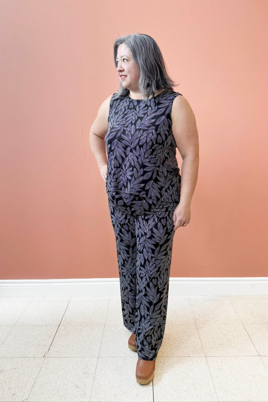 Christina is wearing the Swing Cami by Rien ne se Perd in Black and White Foliage, a trapeze cut tank with a hi-low rounded hemline. She is wearing it with the matching Rumba Palazzo pants and standing  in front of a pink wall. 