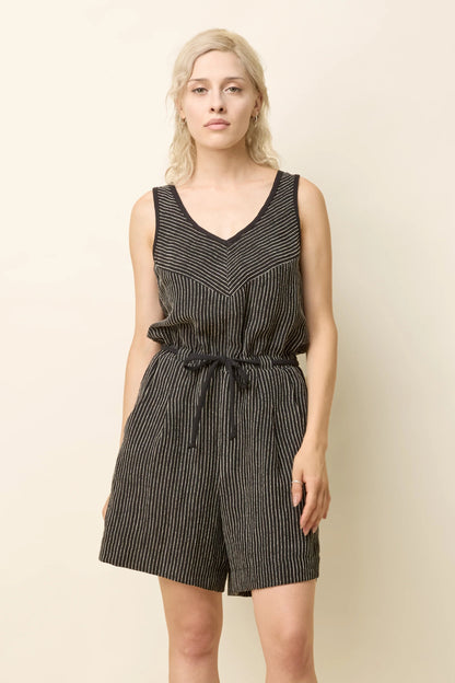 A woman wearing the Shamsa Romper by Cokluch in Black Stripe, a sleeveless romper with a V-neck at the front, a cross-over back, and elastic waist with a tie belt. She is standing in front of a beige background. 