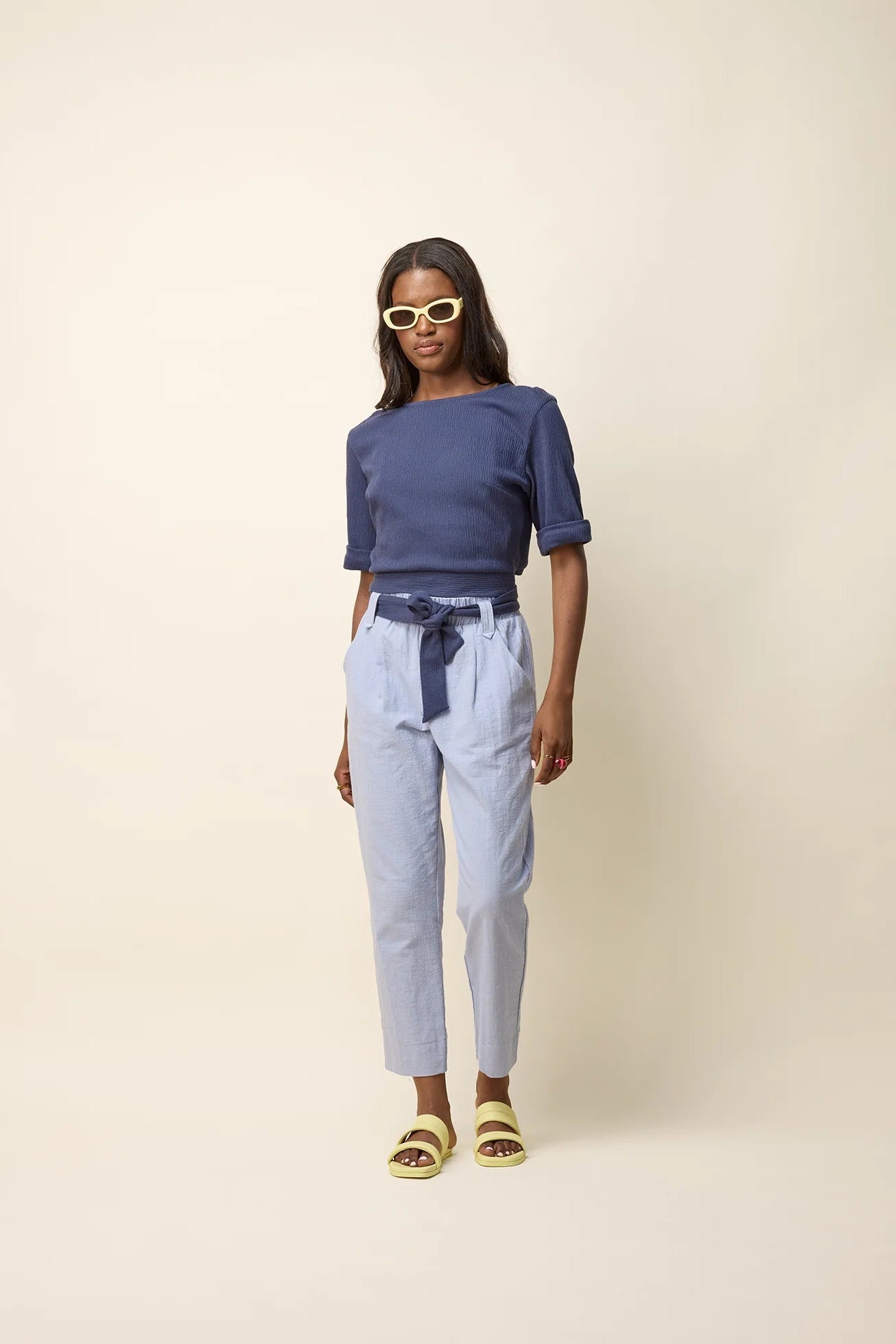 A woman wearing the Jess Top by Cokluch in Navy, a fitted top with a round neck, cuffed elbow-length sleeves, a cut out at the bottom back, and a tie belt. She is wearing it with light blue pants and standing in front of a beige background. 