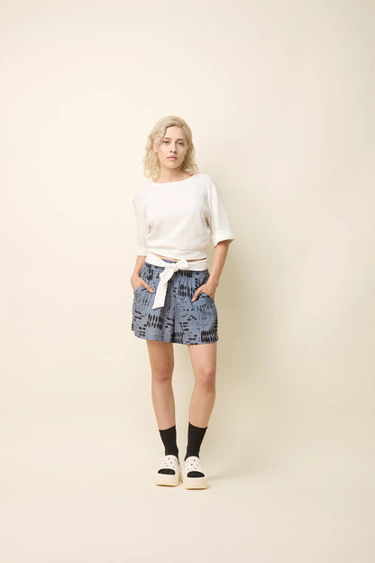 A woman wearing the Jess Top by Cokluch in Off-White, a fitted top with a round neck, cuffed elbow-length sleeves, a cut out at the bottom back, and a tie belt. She is wearing it with the Joy Shorts in Blue Ink and standing in front of a beige background. 